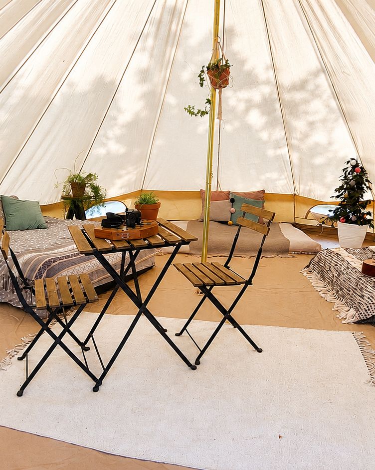 Inside the bell tent, with a double bed made up, a rug, a folding table and chairs, cushions and potted plants around the centre pole