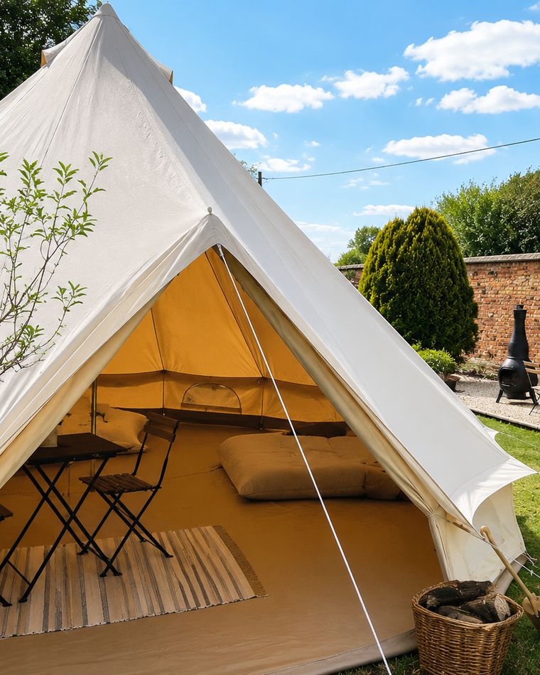 The bell tent pitched on a lawn in a walled garden, doors open, with a table and chairs inside and a log basket by the entrance